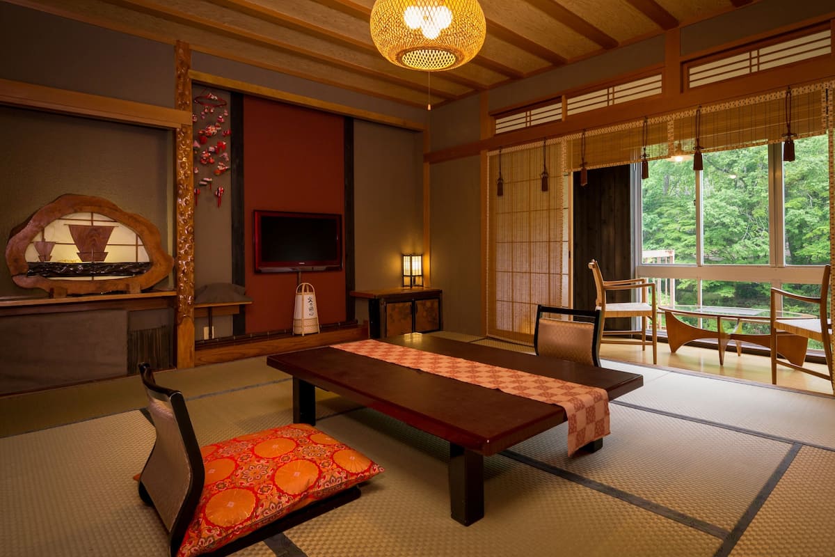 Um quarto japonês tradicional com tatames, uma mesa baixa, cadeiras, televisão e vista para o verde através de grandes janelas.