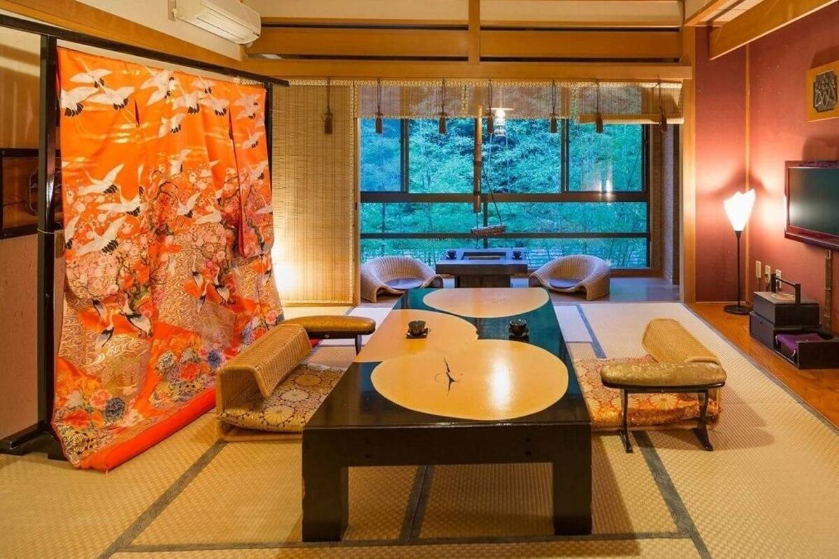 Um quarto japonês tradicional com piso de tatame, uma mesa baixa, almofadas, uma janela grande com vista para árvores e uma televisão.