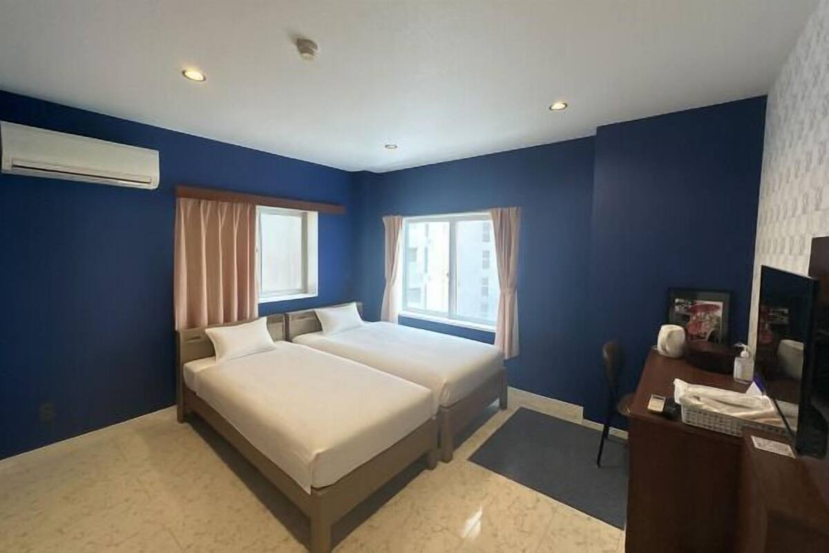 A hotel room with two beds, a desk, a chair, an air conditioning unit, and a window with curtains.