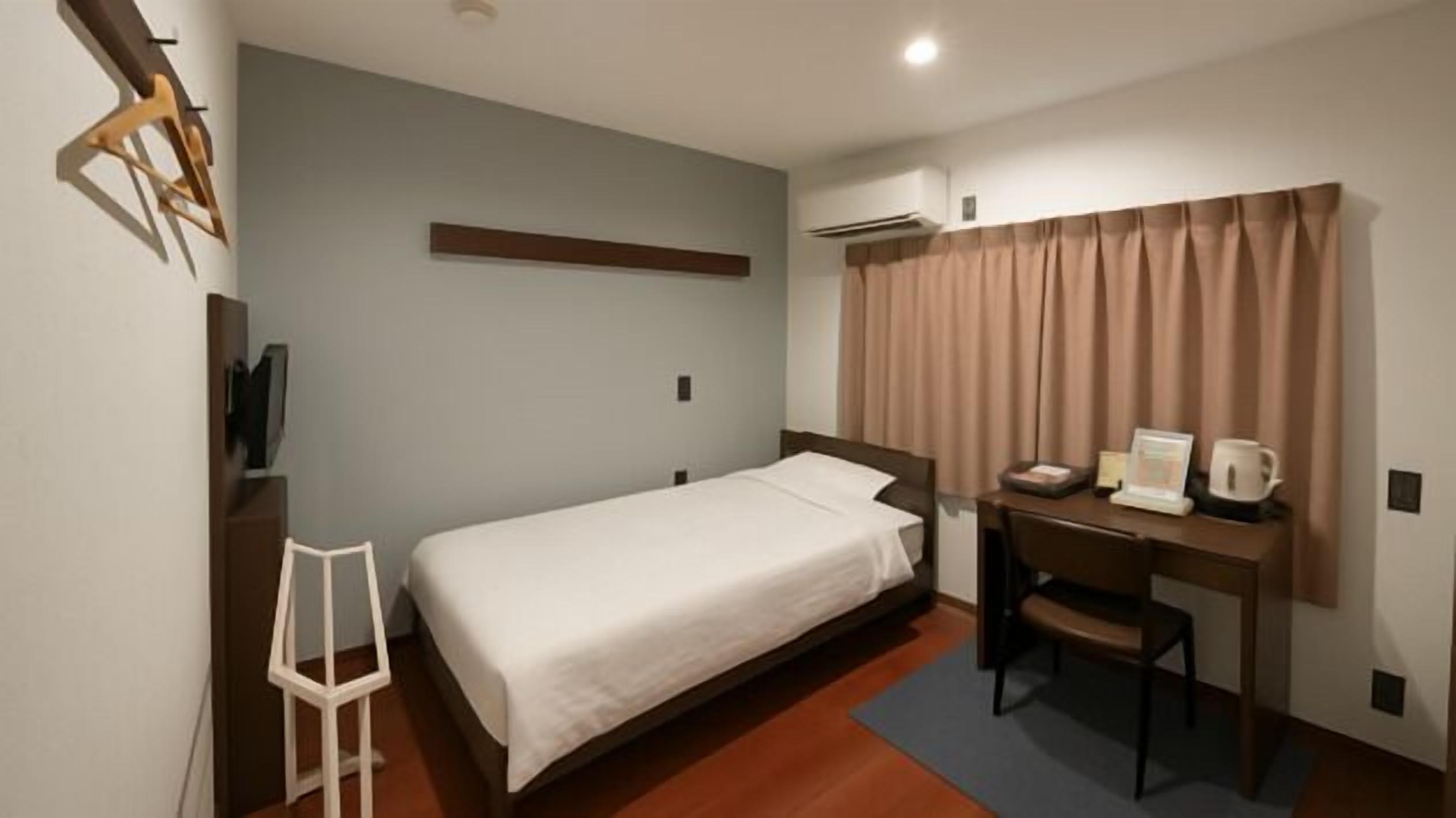 A hotel room with a single bed, a desk with a chair, a small table with a kettle, and a window with curtains.