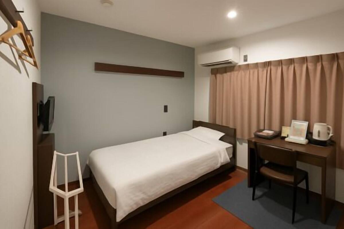 A hotel room with a single bed, a desk with a chair, a small table with a kettle, and a window with curtains.