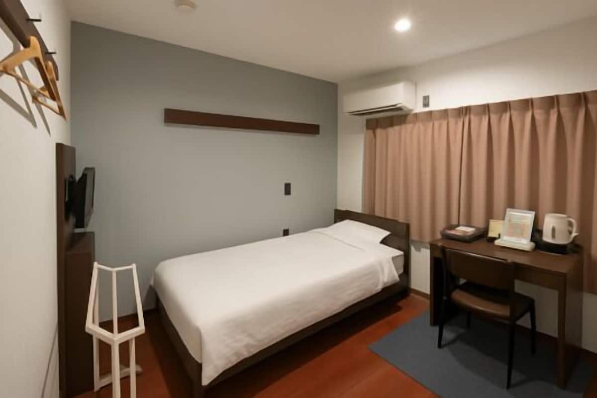 A hotel room with a single bed, a desk with a chair, a small table with a kettle, and a window with curtains.
