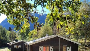 Two wooden cabins with white shutters, surrounded by trees and a mountain backdrop.