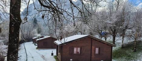 A row of wooden cabins with snow-covered roofs, surrounded by trees and mountains.