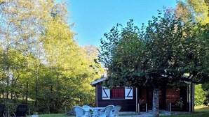 A small building with a dark exterior, a window with red curtains, and a door. There is a white plastic table with chairs in front of it, and a tree to the right. - "Sédour" Chalet de Montorgueil (Saurat)