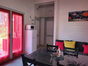 A room with a table, chairs, a refrigerator, a window with red curtains, and a painting on the wall.