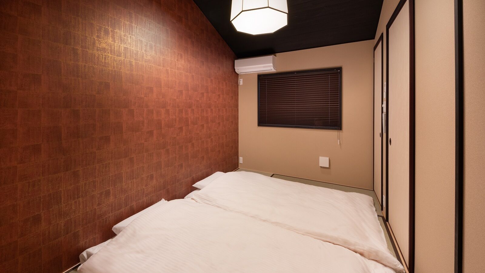 A small, single-bed room with a brick wall, a window with blinds, and a ceiling light fixture.