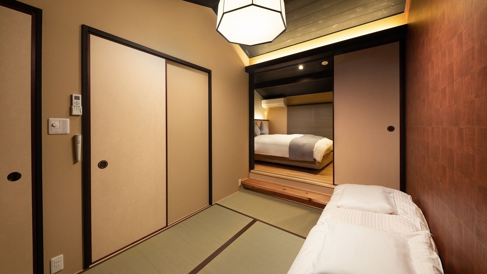 A hotel room with a bed, a mirror, a bedside table, a wall-mounted heater, and a ceiling light fixture.