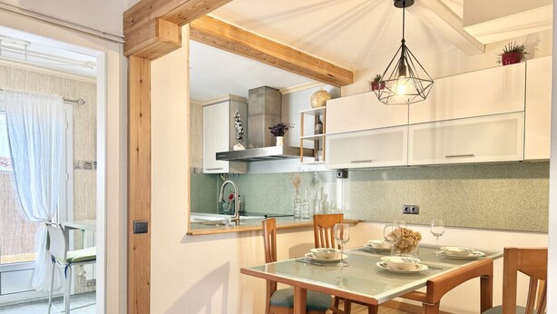 Un coin repas avec une table en verre, des chaises en bois, une cuisine en arrière-plan et un luminaire au plafond.