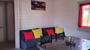 A room with a white wooden wall, a blue sofa with red and yellow cushions, a dining table with a plastic bag of biscuits, and a painting above the sofa.