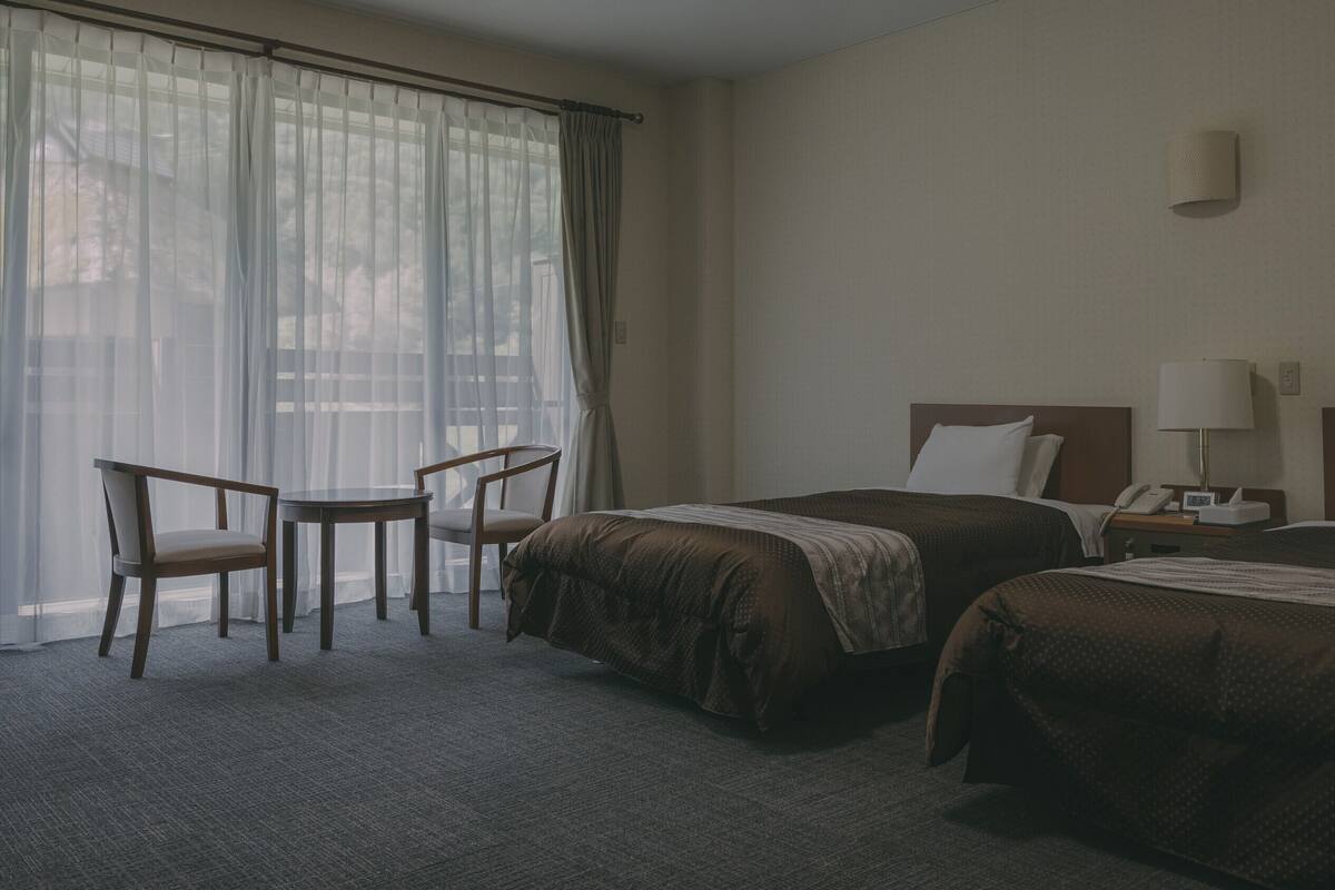 Habitación de hotel con dos camas, una mesita pequeña, una silla, una mesita de noche con lámpara y una ventana con cortinas translúcidas.