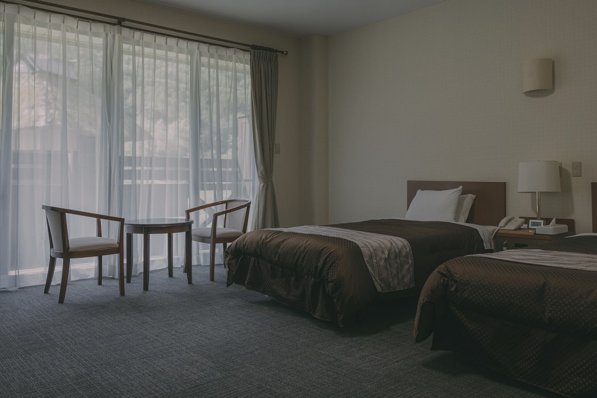 Une chambre d’hôtel avec deux lits, une petite table, une chaise, une table de chevet avec une lampe et une fenêtre avec des rideaux légers.