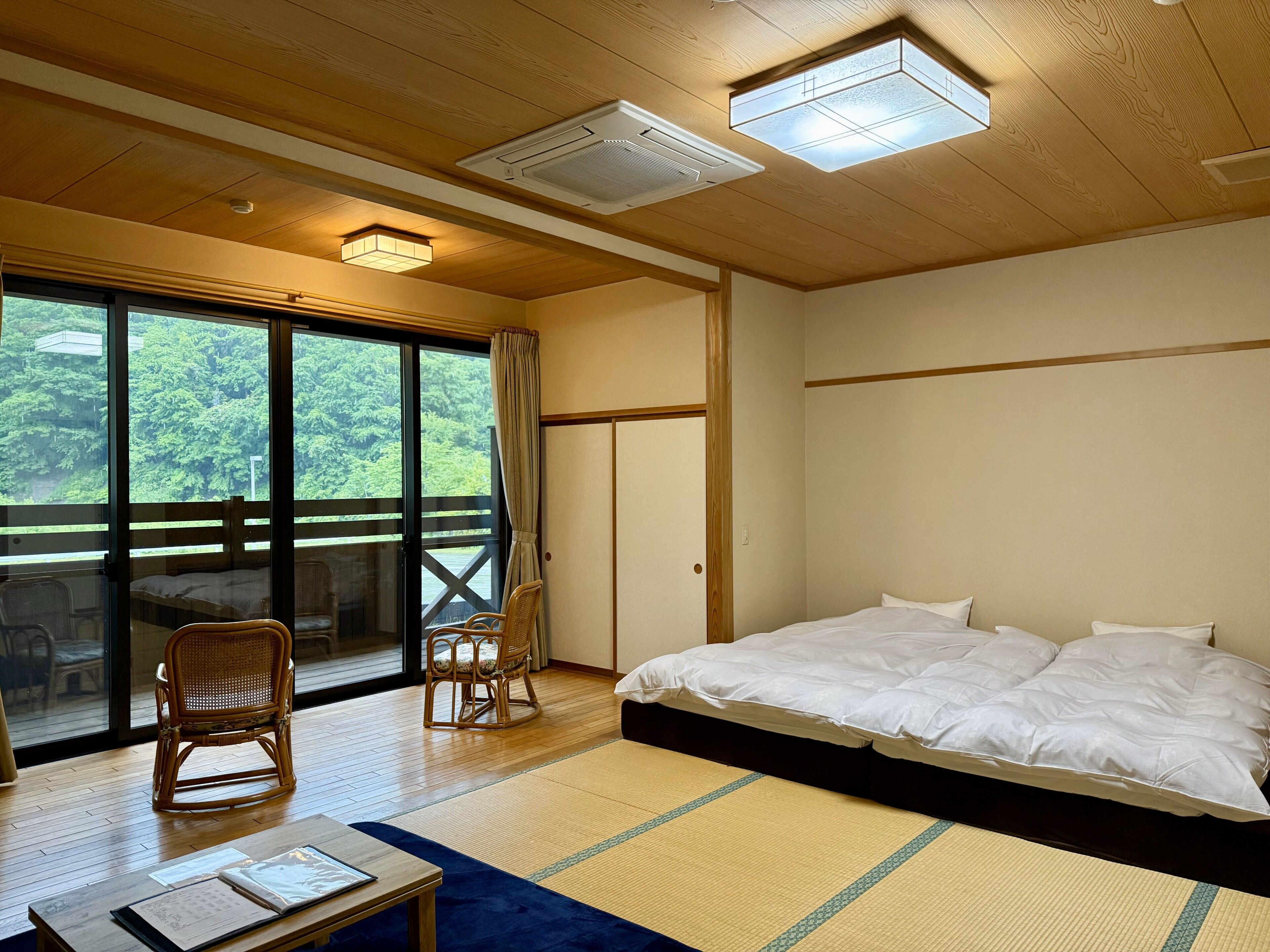 A room with a bed, two chairs, a table, and a view of greenery through large windows.