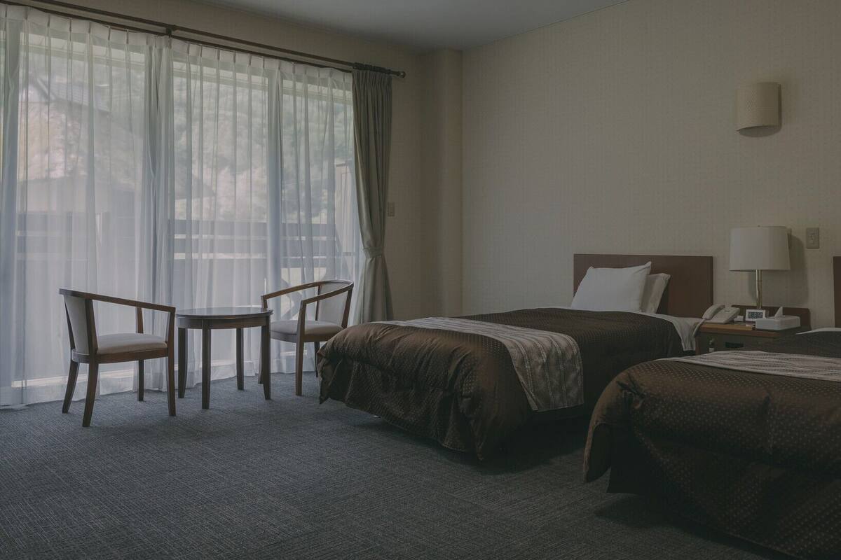 Habitación de hotel con dos camas, una mesita, una silla y una ventana con cortinas de gasa.