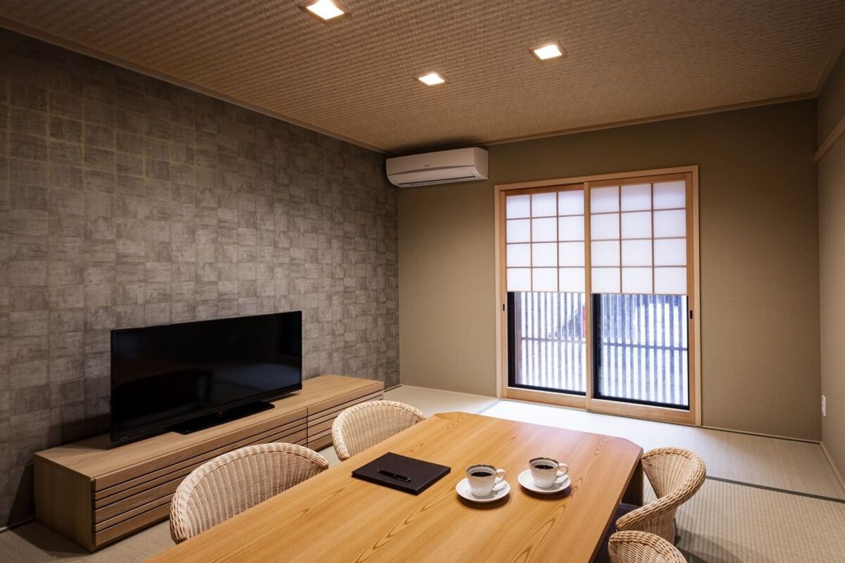 A room with a TV, a wooden table with chairs, a sliding door, and a view through the door.