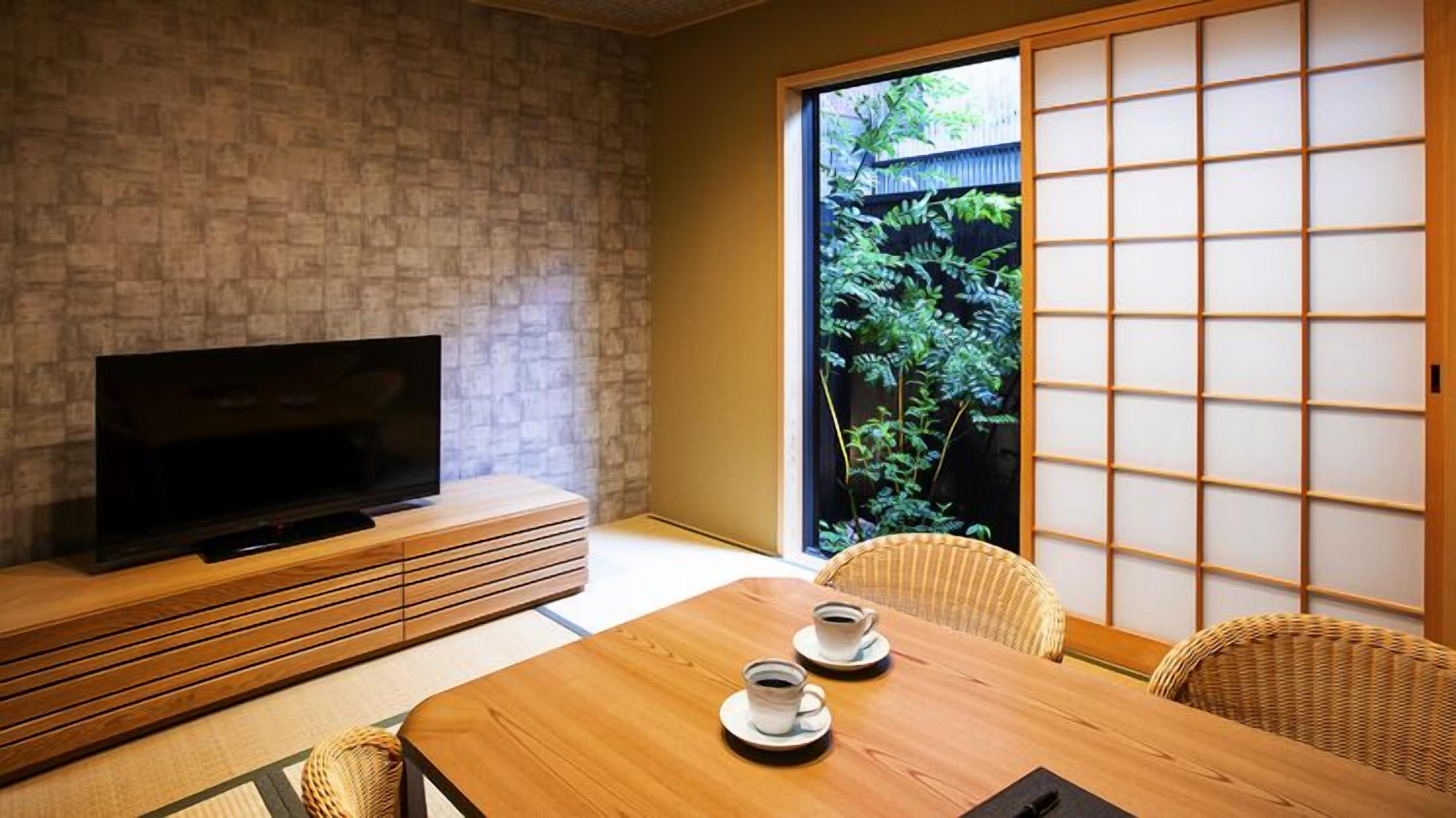 A traditional Japanese room with tatami flooring, a television on a low wooden stand, a sliding door with a view of greenery, and a table with two cups.