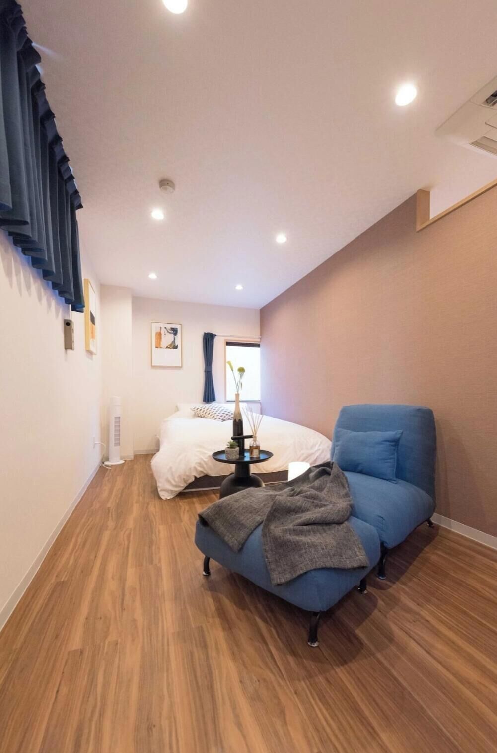 A modern hotel room with a wooden floor, a blue armchair, a bed with white bedding, and a small table with a vase.