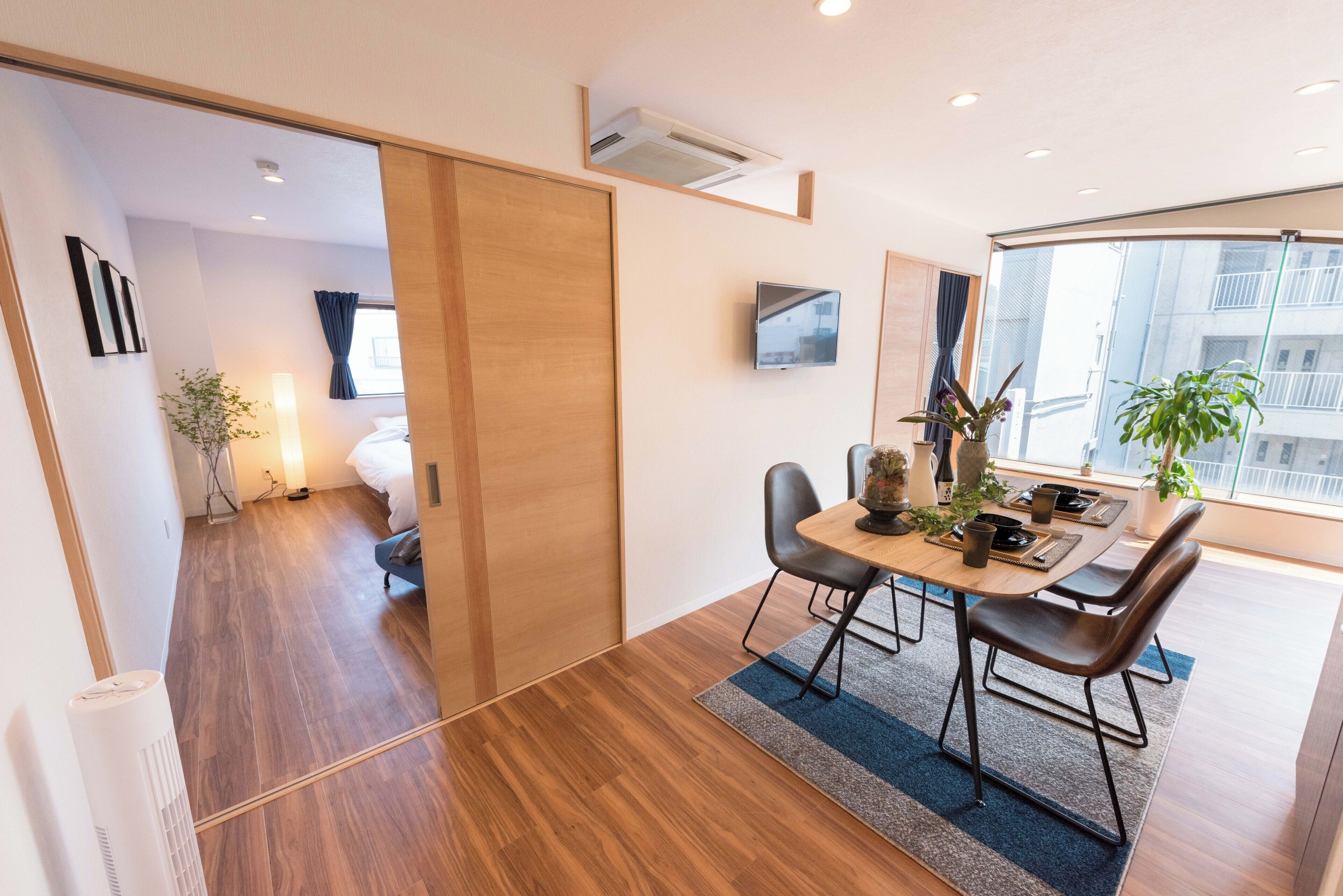 Una moderna camera d'albergo con pavimento in legno, una zona pranzo con tavolo e sedie, una TV fissata al muro e un'ampia finestra con vista sulla città.