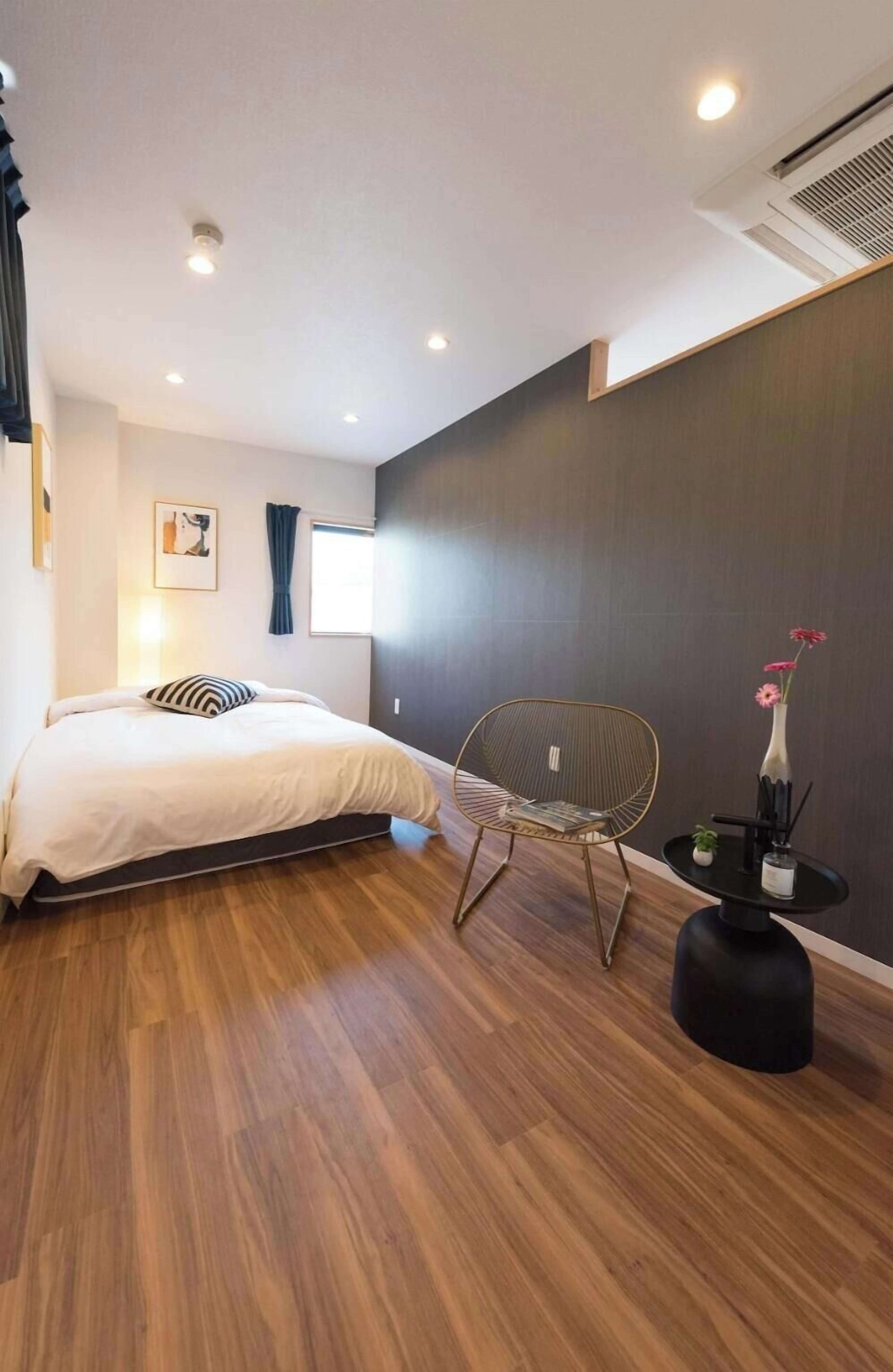 A modern bedroom with a wooden floor, a bed with a white duvet, a chair, a small table with a vase, and a wall-mounted air conditioner.