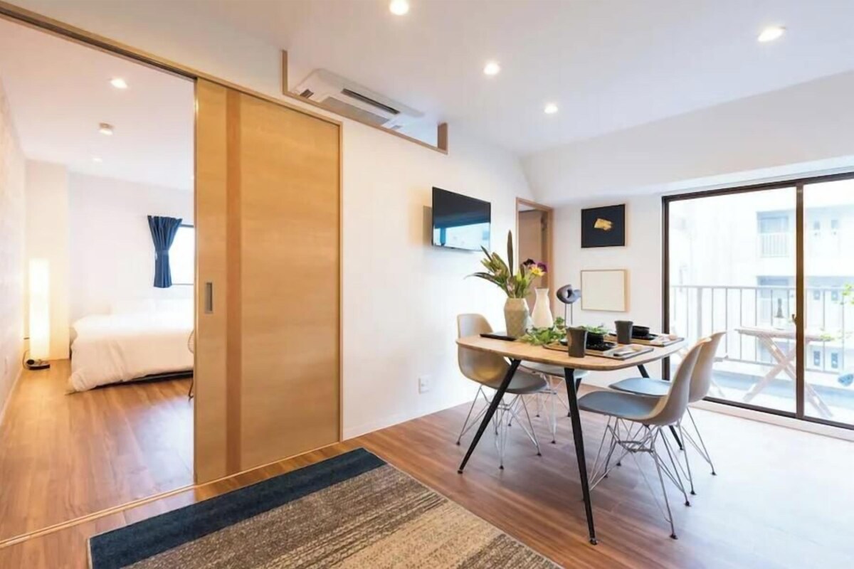 A modern hotel room with a wooden floor, a bed, a desk with chairs, a TV, and a balcony with a view.