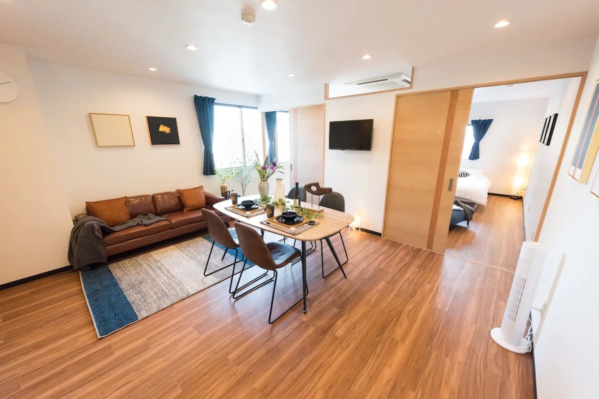 A modern living room with a brown leather sofa, a dining table with chairs, and a large window with blue curtains.