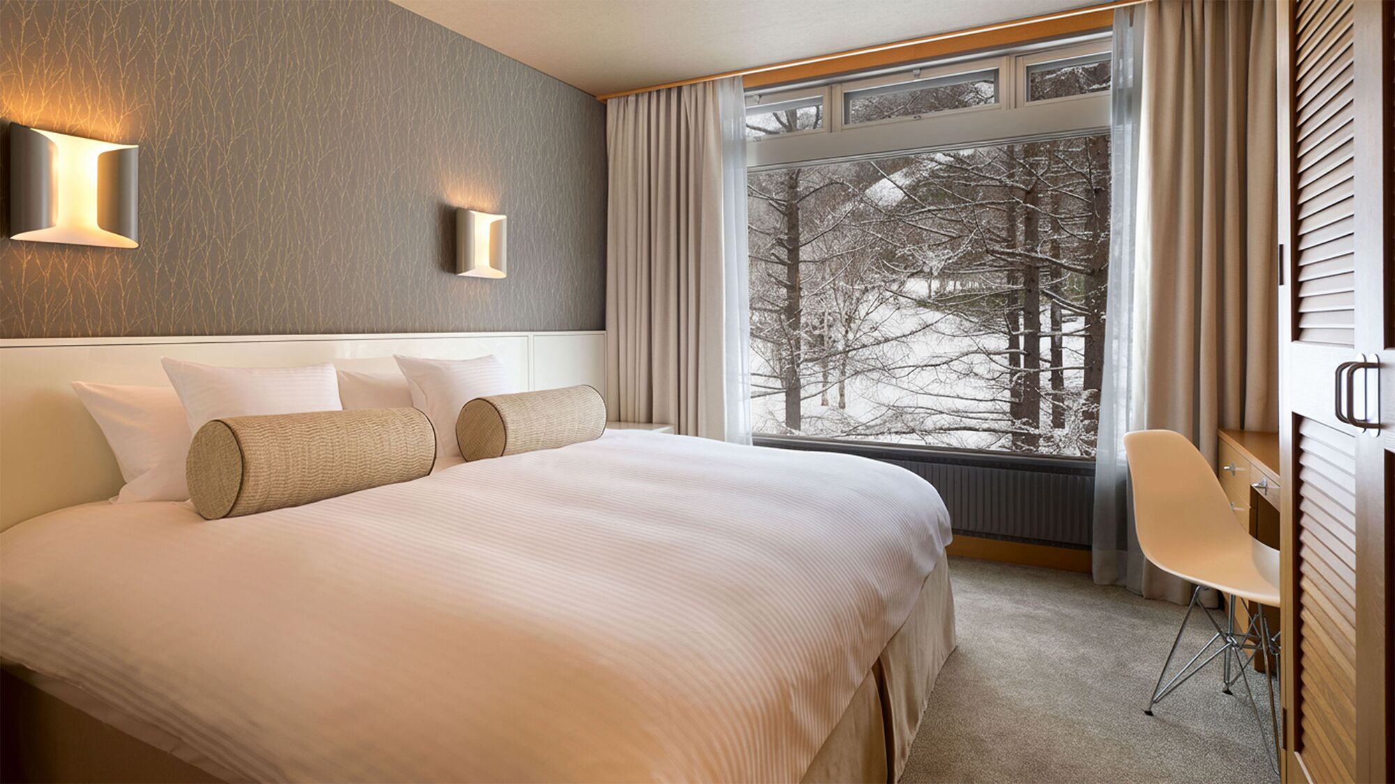 Ein Hotelzimmer mit einem großen Bett, zwei Wandleuchten, Blick auf Bäume durch das Fenster und einem Schreibtisch mit Stuhl.