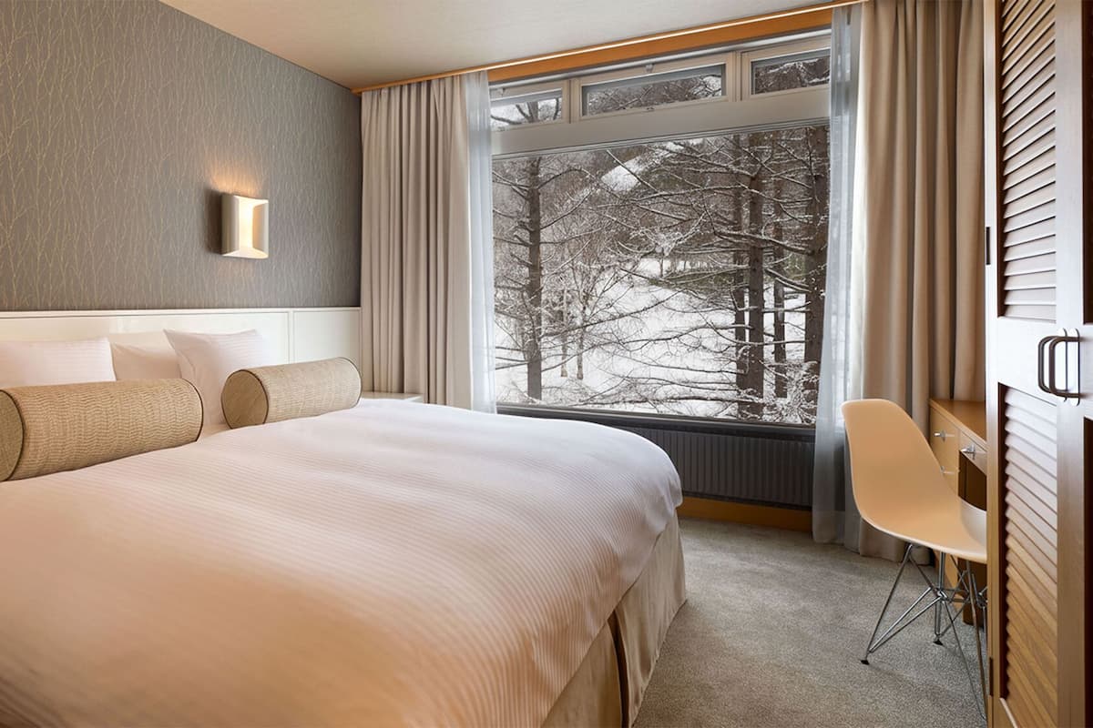 A hotel room with a large bed, two wall-mounted lights, a view of trees through the window, and a desk with a chair.