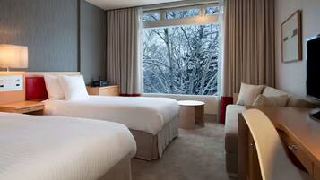 A hotel room with two beds, a desk, a chair, a TV, and a window with a view of trees.