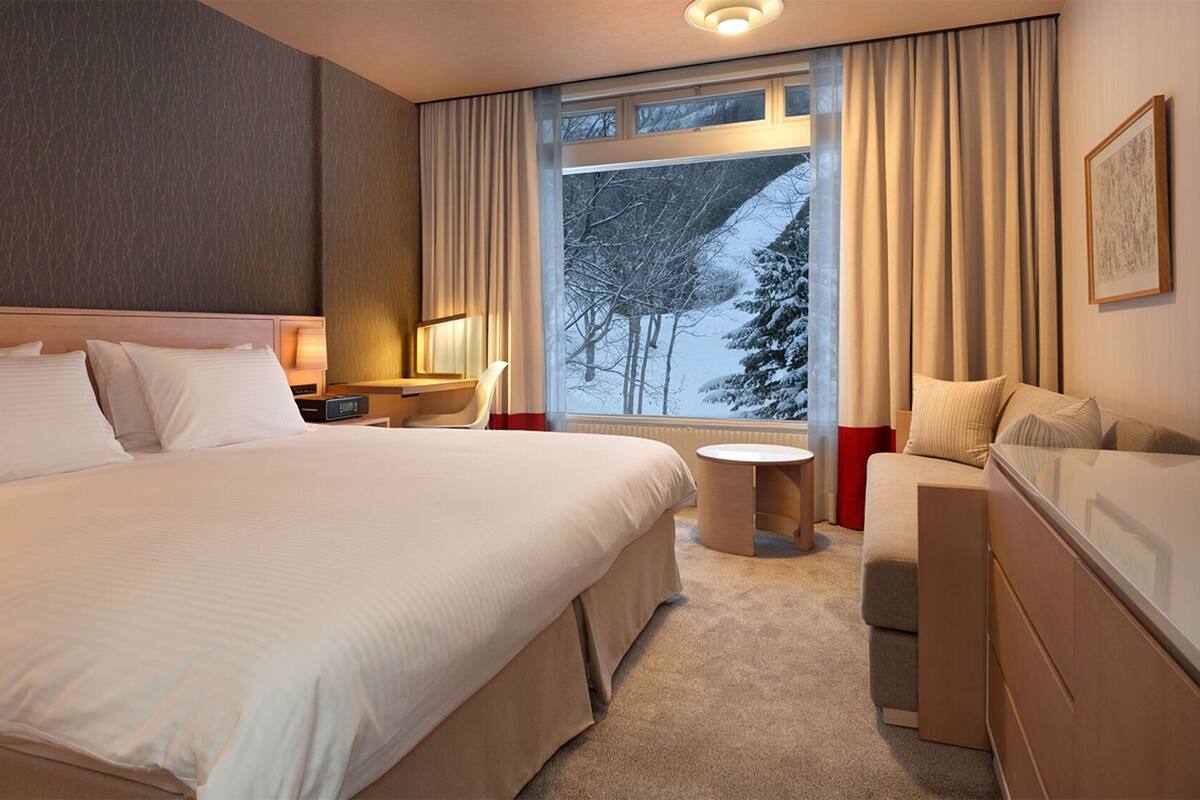 Habitación de hotel con una cama grande, un escritorio, una silla, una mesita y una ventana con vistas a árboles.