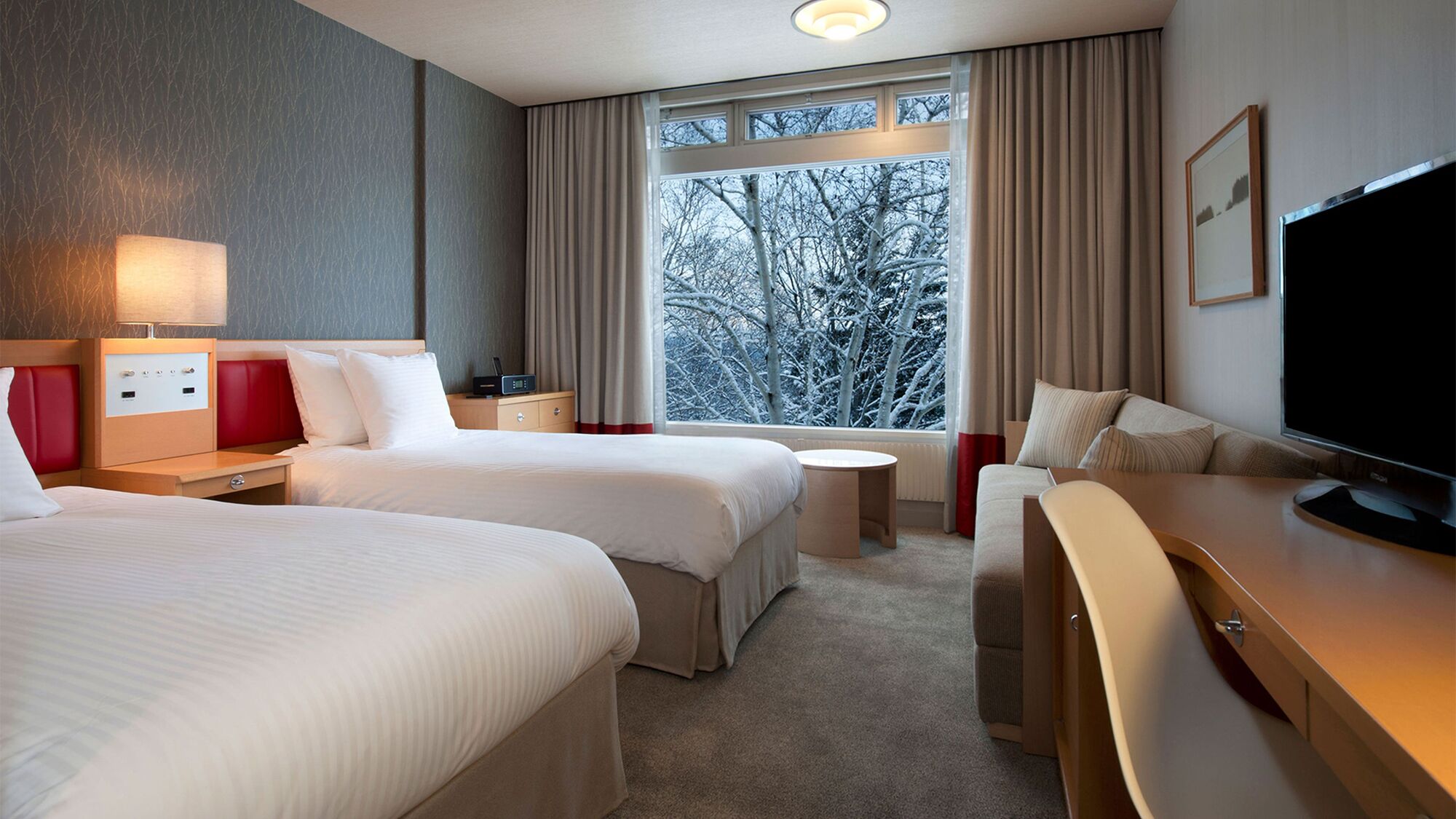 Ein Hotelzimmer mit zwei Betten, einem Fernseher, einem Schreibtisch und einem Fenster mit Blick auf Bäume.