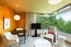 A modern hotel room with a large window, a flat-screen TV, a dining table, and two chairs.