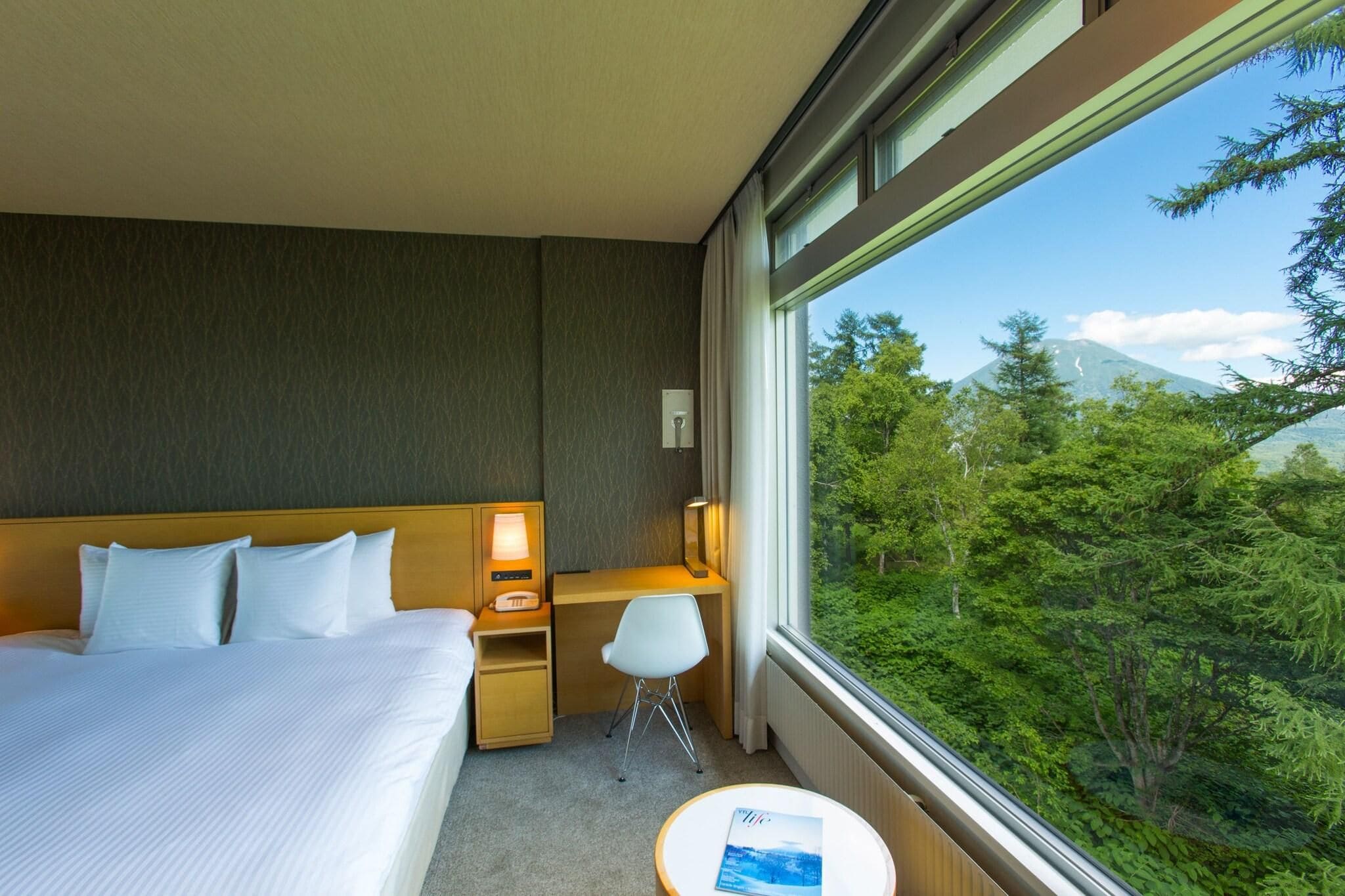 Habitación de hotel con cama, escritorio, silla y vistas a un bosque y una montaña.