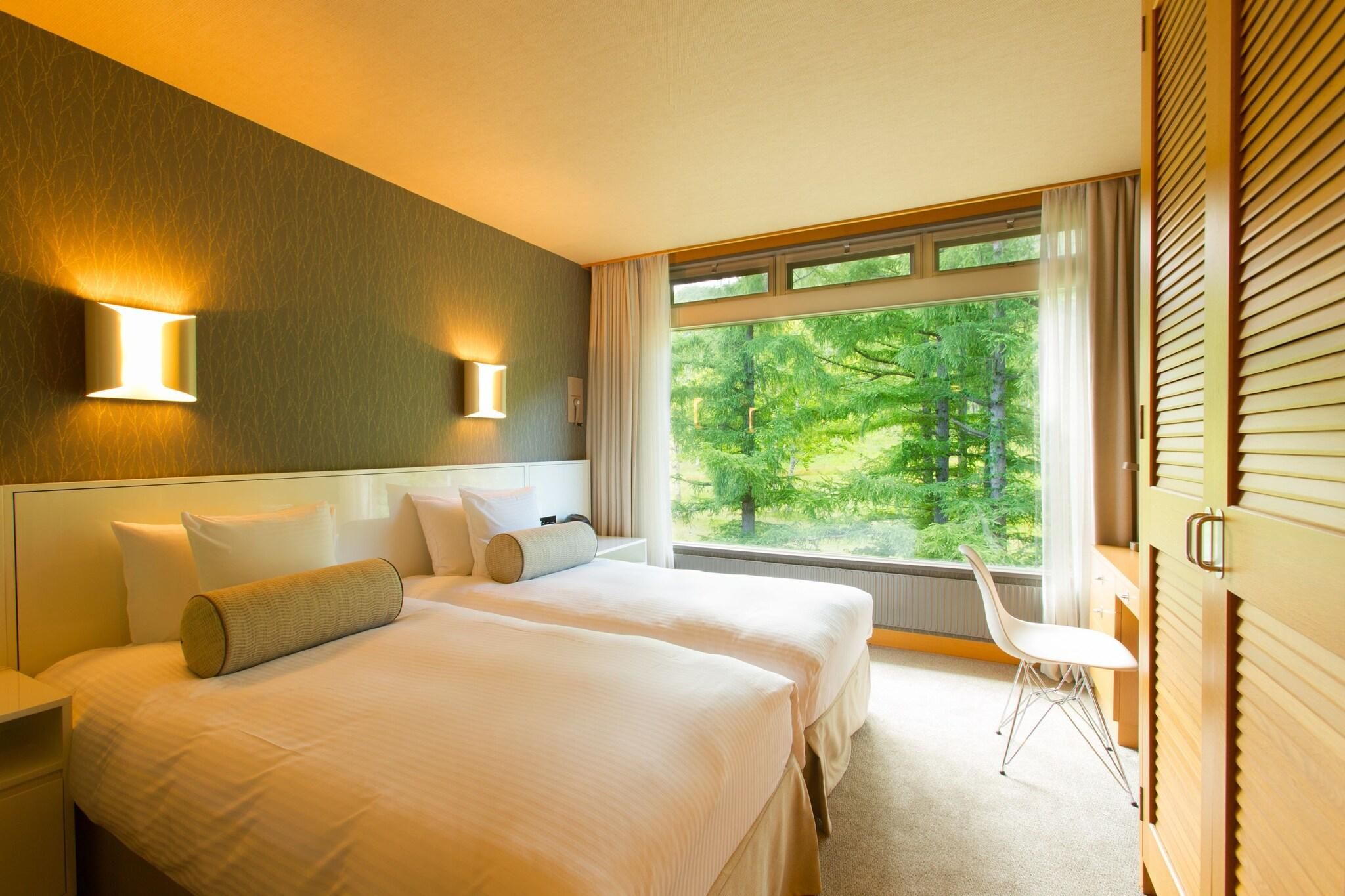 A hotel room with two beds, a window with a view of greenery, and a wall-mounted light fixture.
