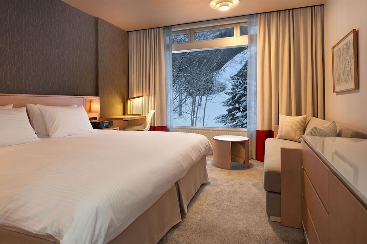A hotel room with a large bed, a desk, a chair, a small table, and a window with a view of trees.
