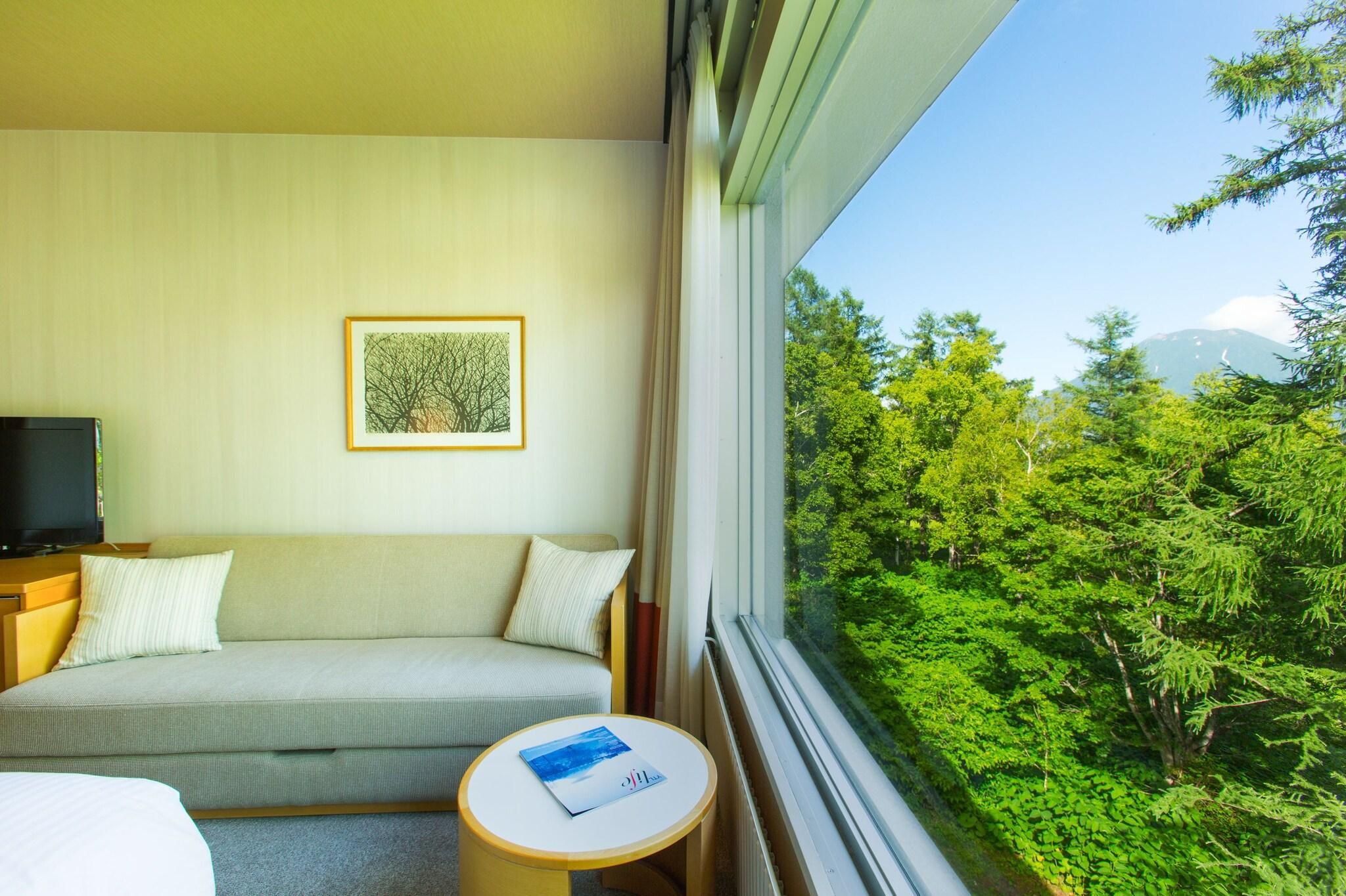 A hotel room with a sofa, a small table, a TV, and a window with a view of greenery and mountains.