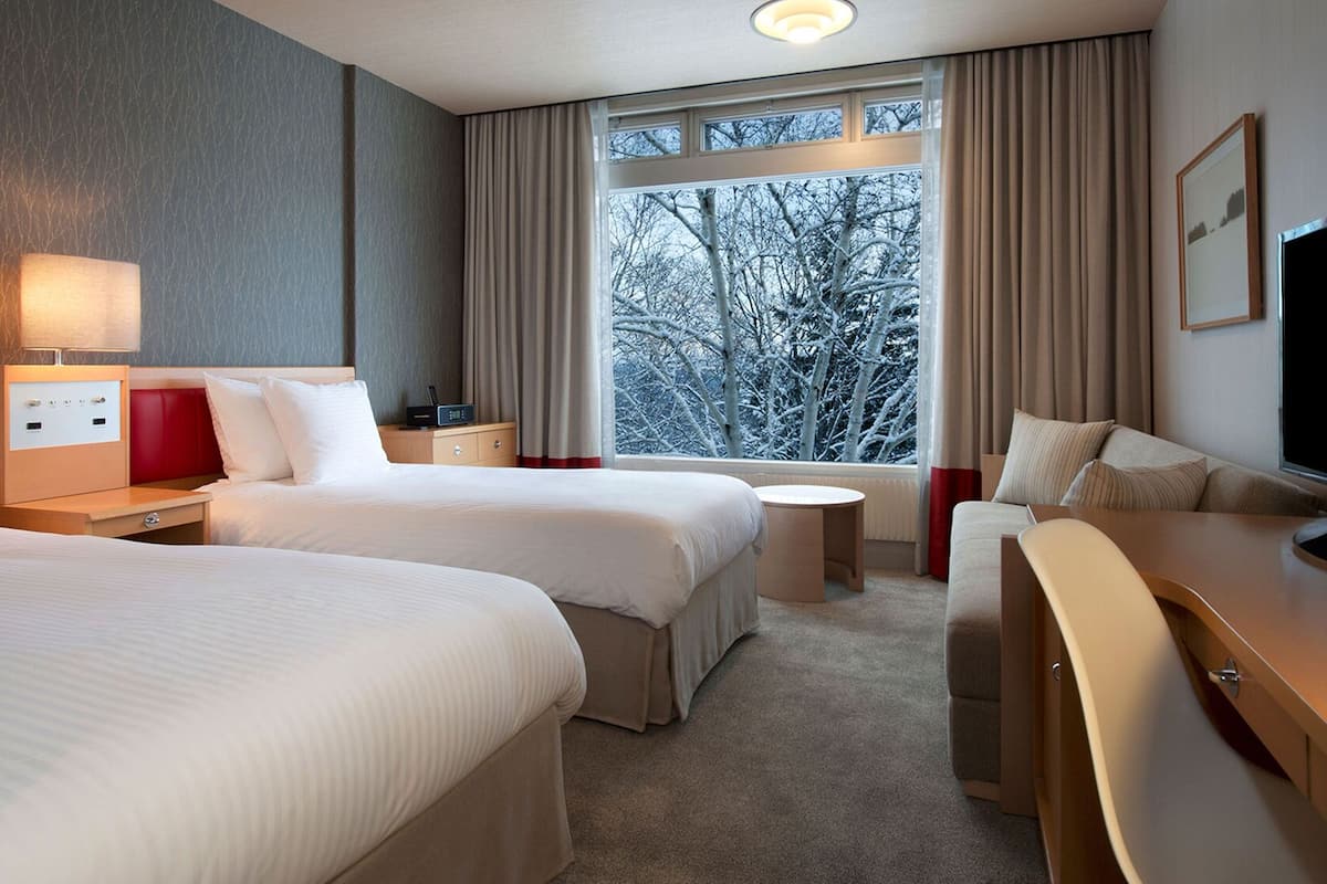 A hotel room with two beds, a TV, a desk, and a window with a view of trees.