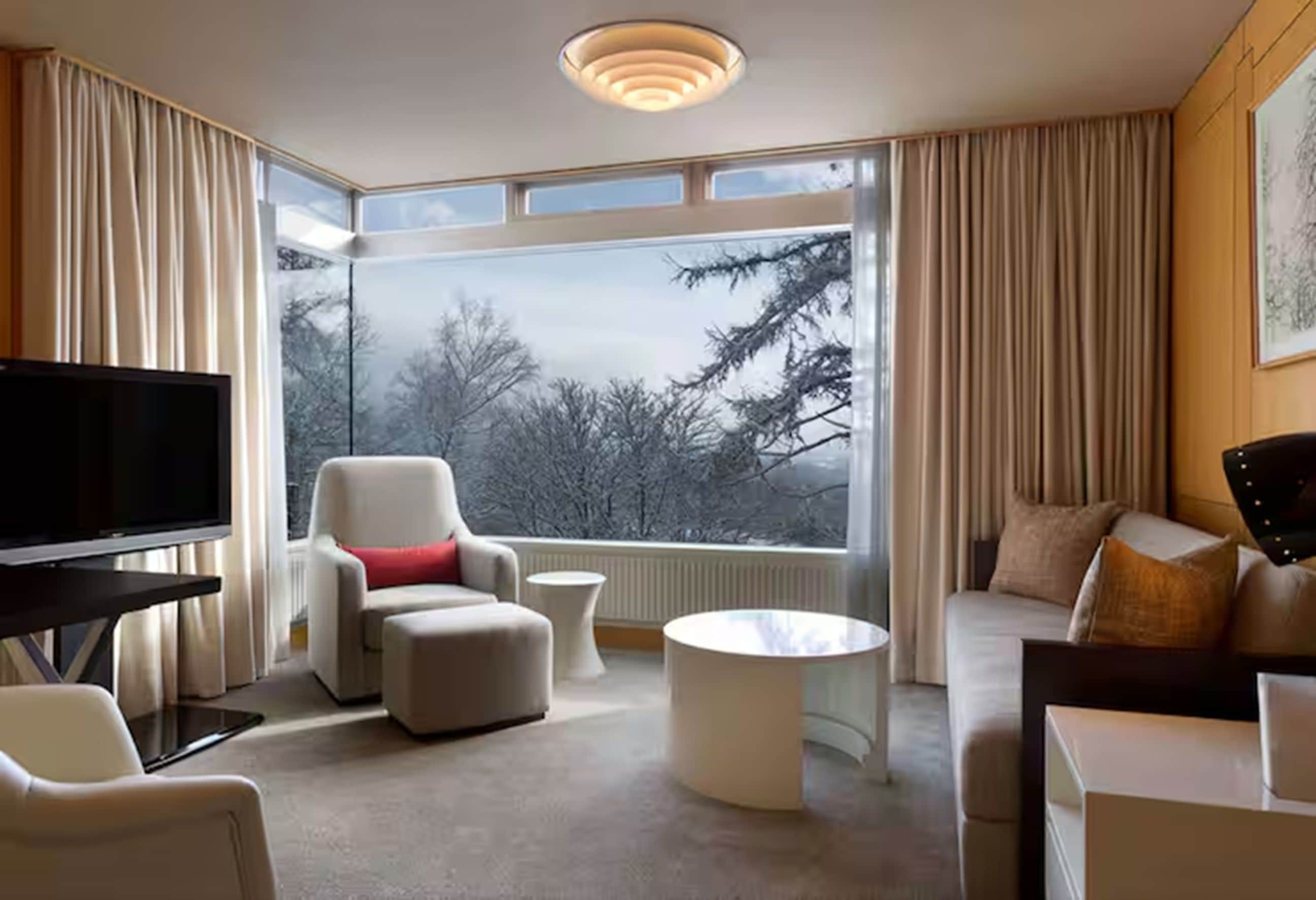 Una habitación de hotel moderna con un ventanal grande, un televisor de pantalla plana, un sofá, una silla y una mesa de centro.