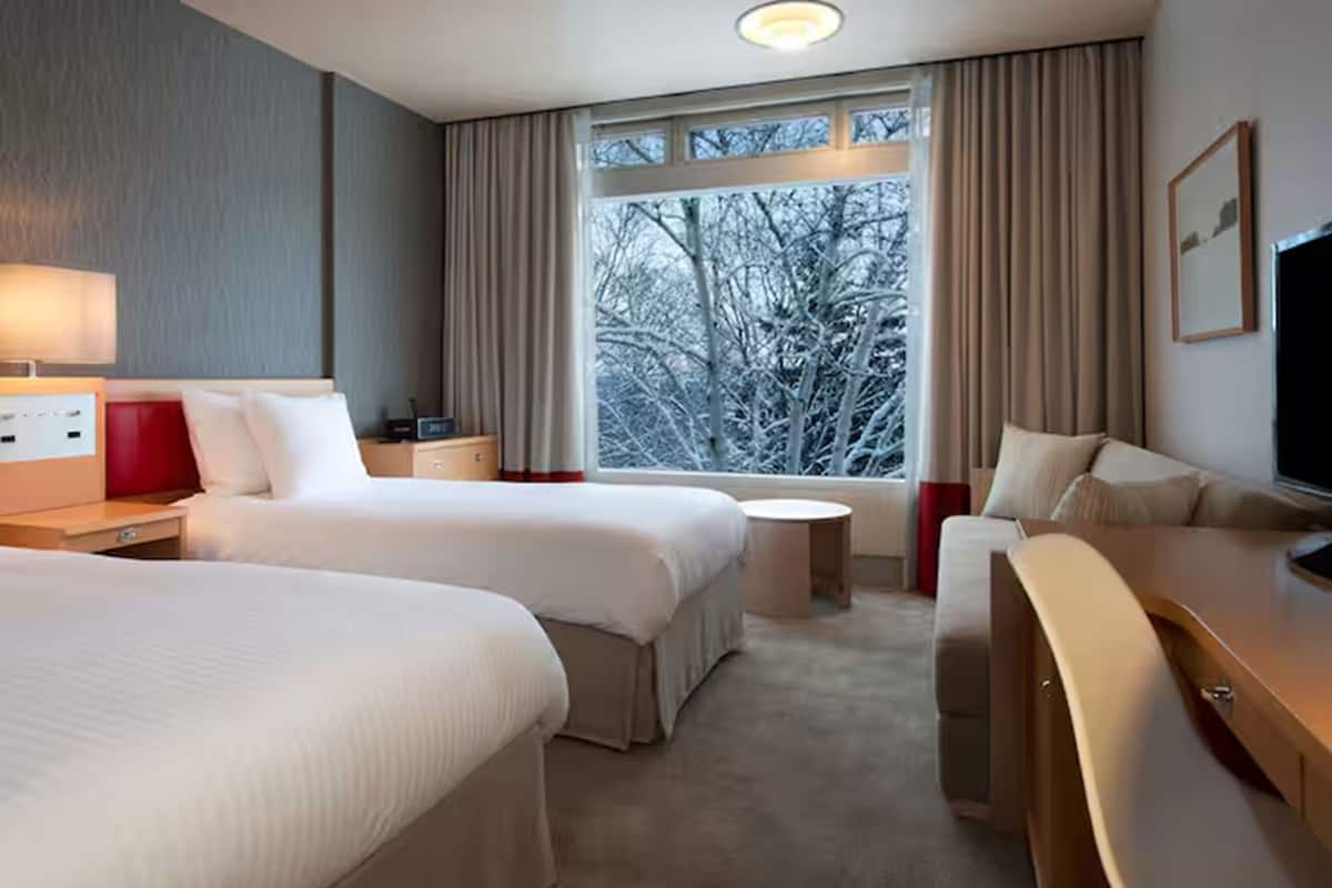 A hotel room with two beds, a desk, a chair, a TV, and a window with a view of trees.