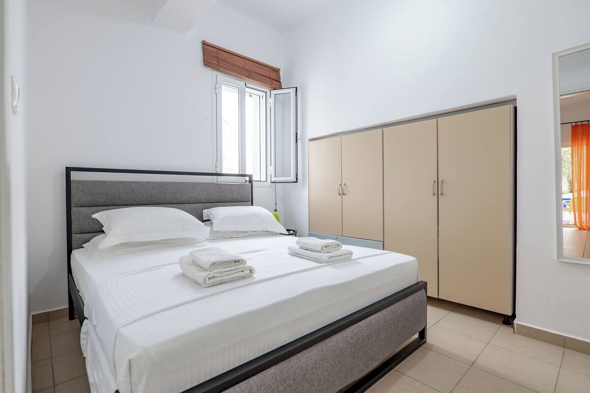 A neatly made bed with pillows and folded towels, a wardrobe, and a window with blinds.