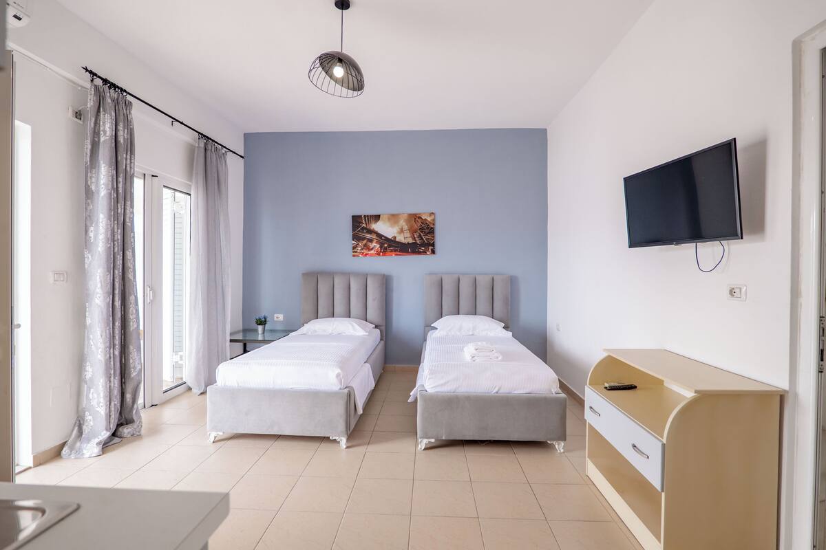A hotel room with two beds, a TV, a desk, and a window with curtains.