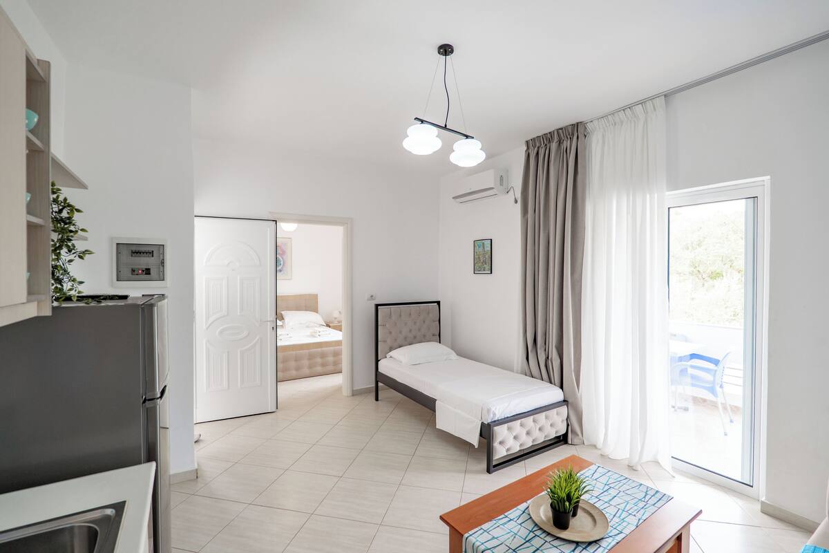 A modern hotel room with a bed, a dining table, a kitchen area, and a sliding door leading to a balcony.