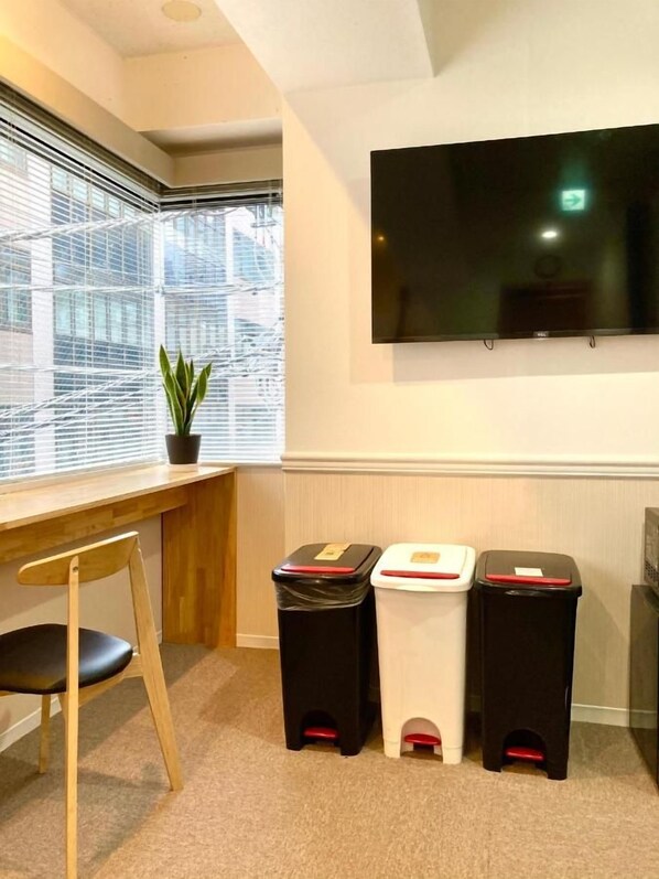 A room with a desk, chair, a potted plant, a TV mounted on the wall, and three trash bins.