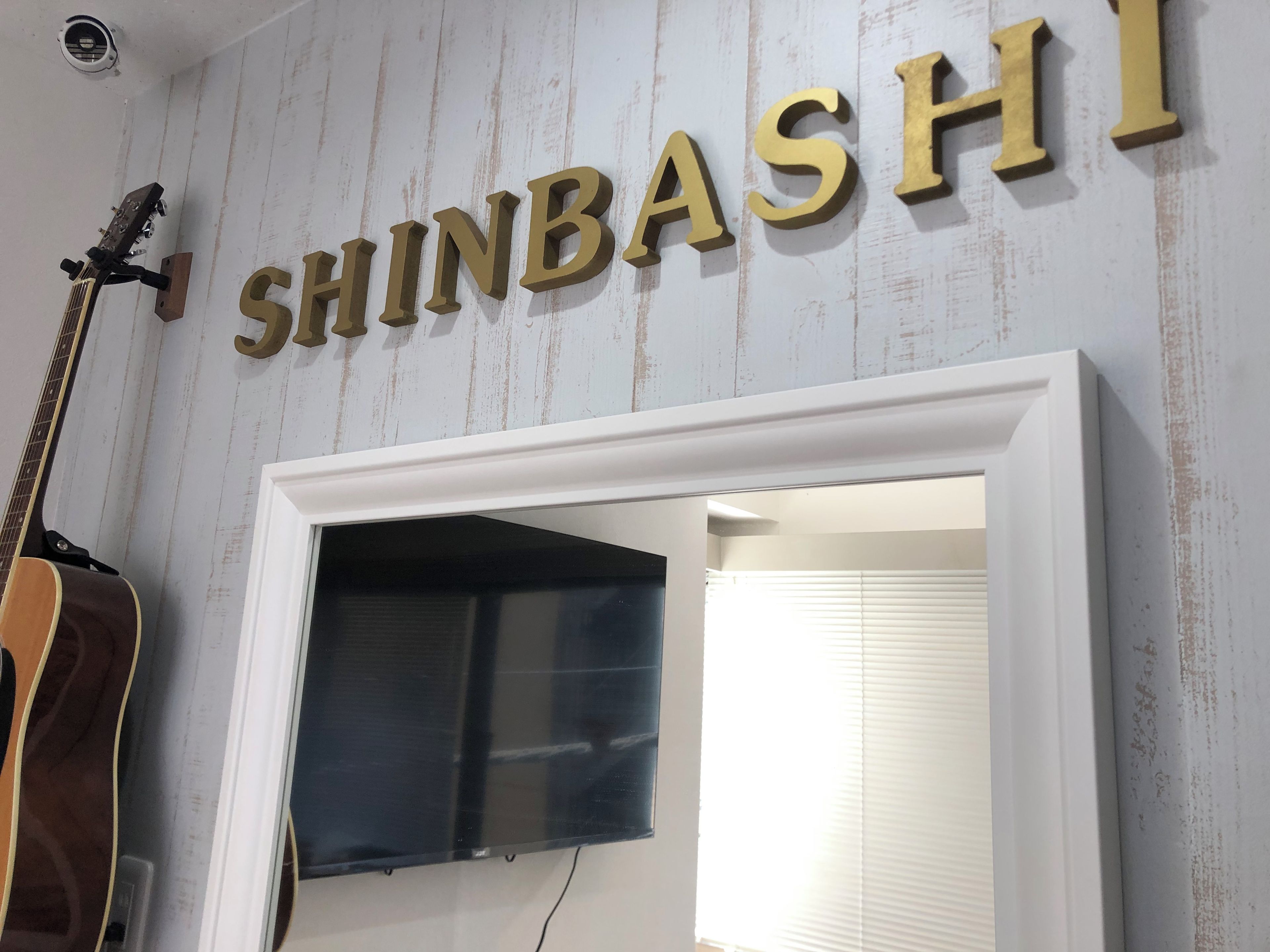 A wall with the name 'SHINBASHI' in large letters, a guitar leaning against the wall, and a doorway leading to another room.