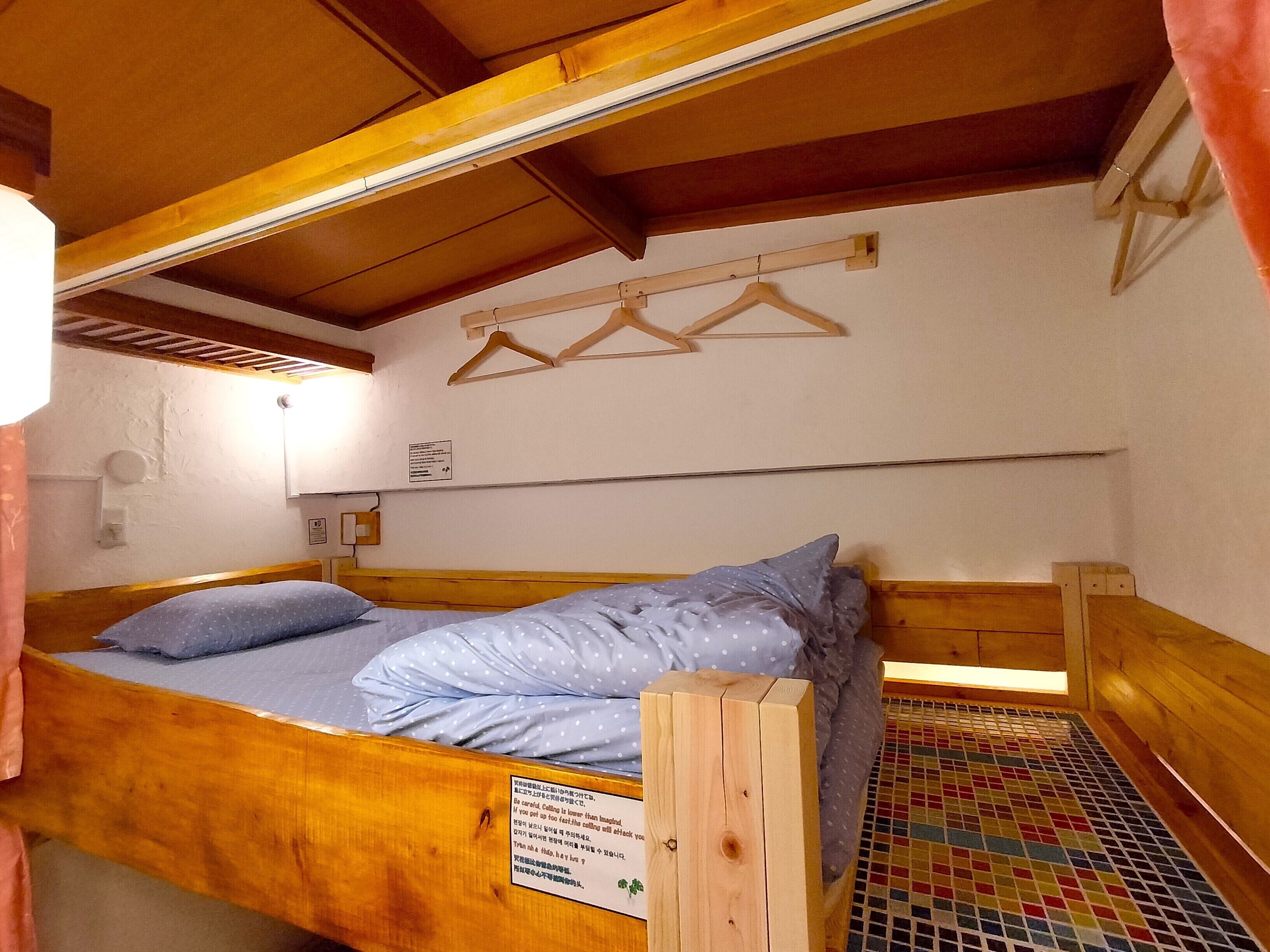 Una cama de madera con colchón y almohadas, cabecera de madera, lámpara de pared y una ventana pequeña con cortina.