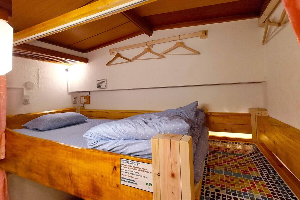 A single wooden bed with a mattress and pillows, a wooden headboard, a wall-mounted light fixture, and a small window with a curtain.