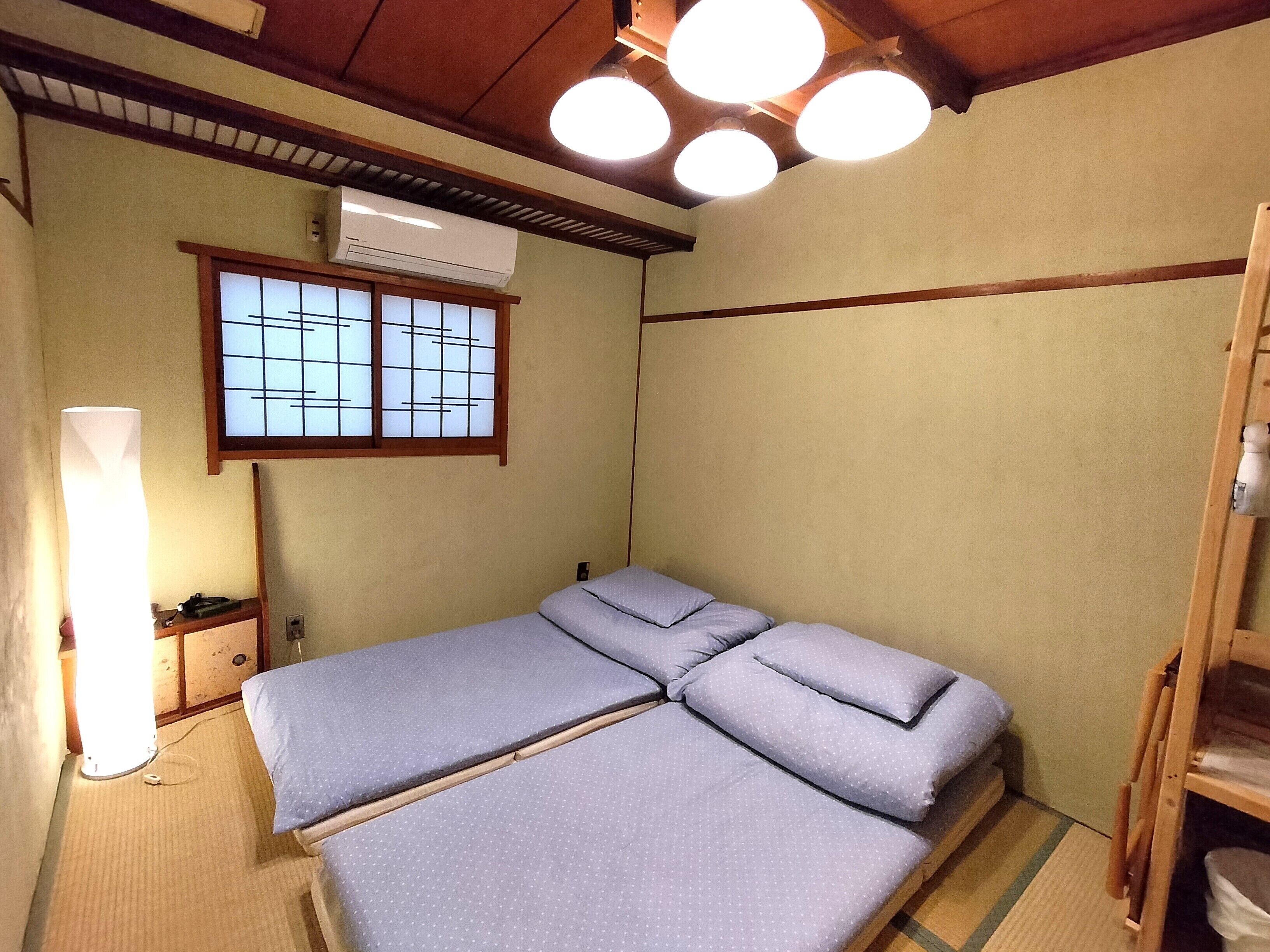 Una habitación tradicional japonesa con suelo de tatami, una cama baja de madera y un panel deslizante, una ventana con celosía de shoji y un techo con múltiples luminarias.