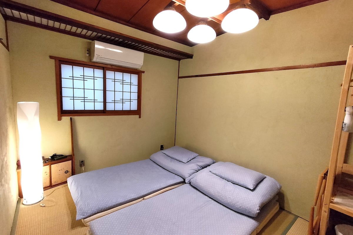 A traditional Japanese-style room with tatami flooring, a low wooden bed with a sliding partition, a shoji screen window, and a ceiling with multiple light fixtures.