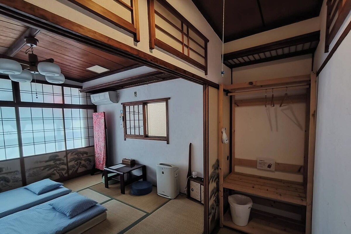 A traditional Japanese-style room with tatami flooring, a futon bed, a small table with a chair, a heater, and a wooden wardrobe with a hanging rack.