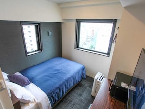 Ein kleines Hotelzimmer mit einem Bett, zwei Fenstern, einer Heizung und einem Schreibtisch mit Computer.