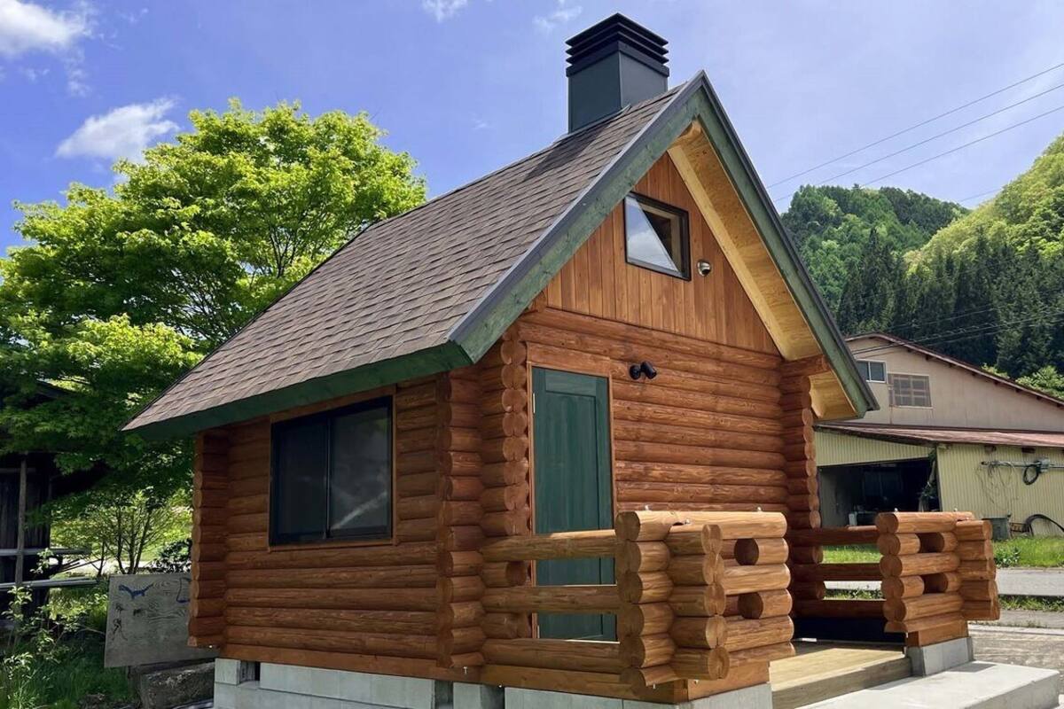 A small log cabin with a pitched roof, a chimney, and a porch with wooden railings.